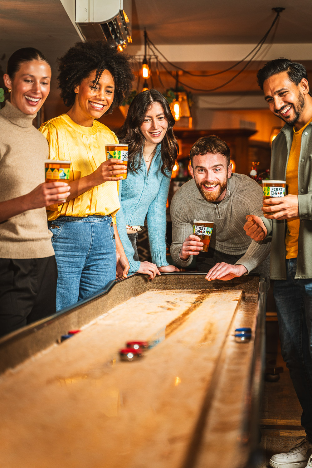 Lifestyle brand image of First Drop beer with friends playing pub games. Group of friends in a pub drinking First Drop beer while playing a shuffleboard-style game