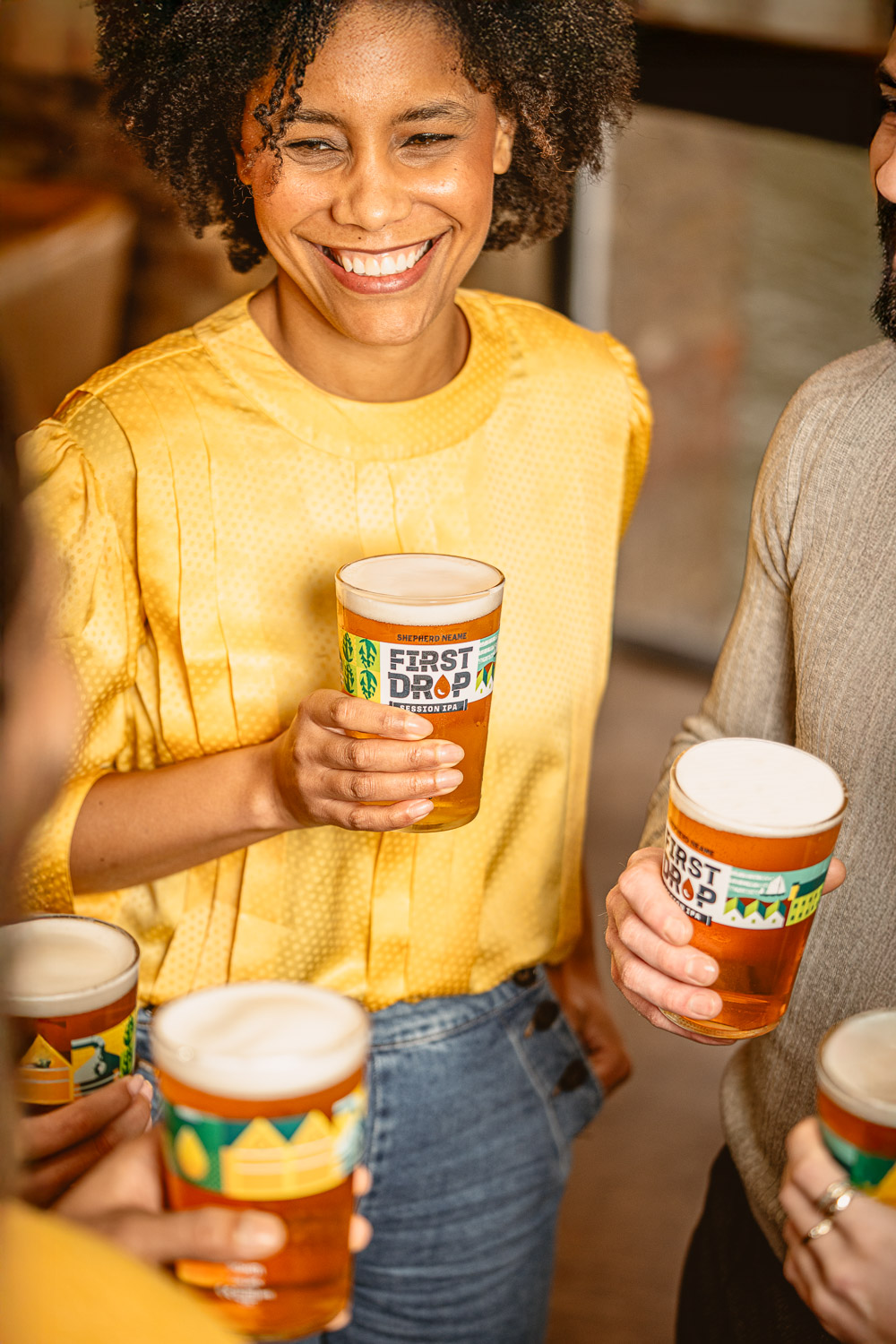 Lifestyle brand photo featuring First Drop beer in a social setting. Smiling woman in yellow holding a pint of First Drop beer with friends at a pub