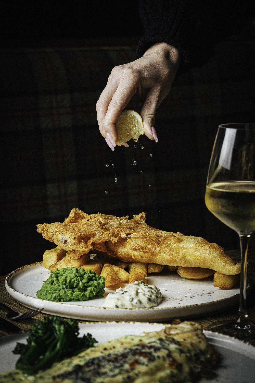 Food styling photo of fish and chips with mushy peas, lemon and wine. Fish and chips with mushy peas, tartar sauce, and a hand squeezing lemon