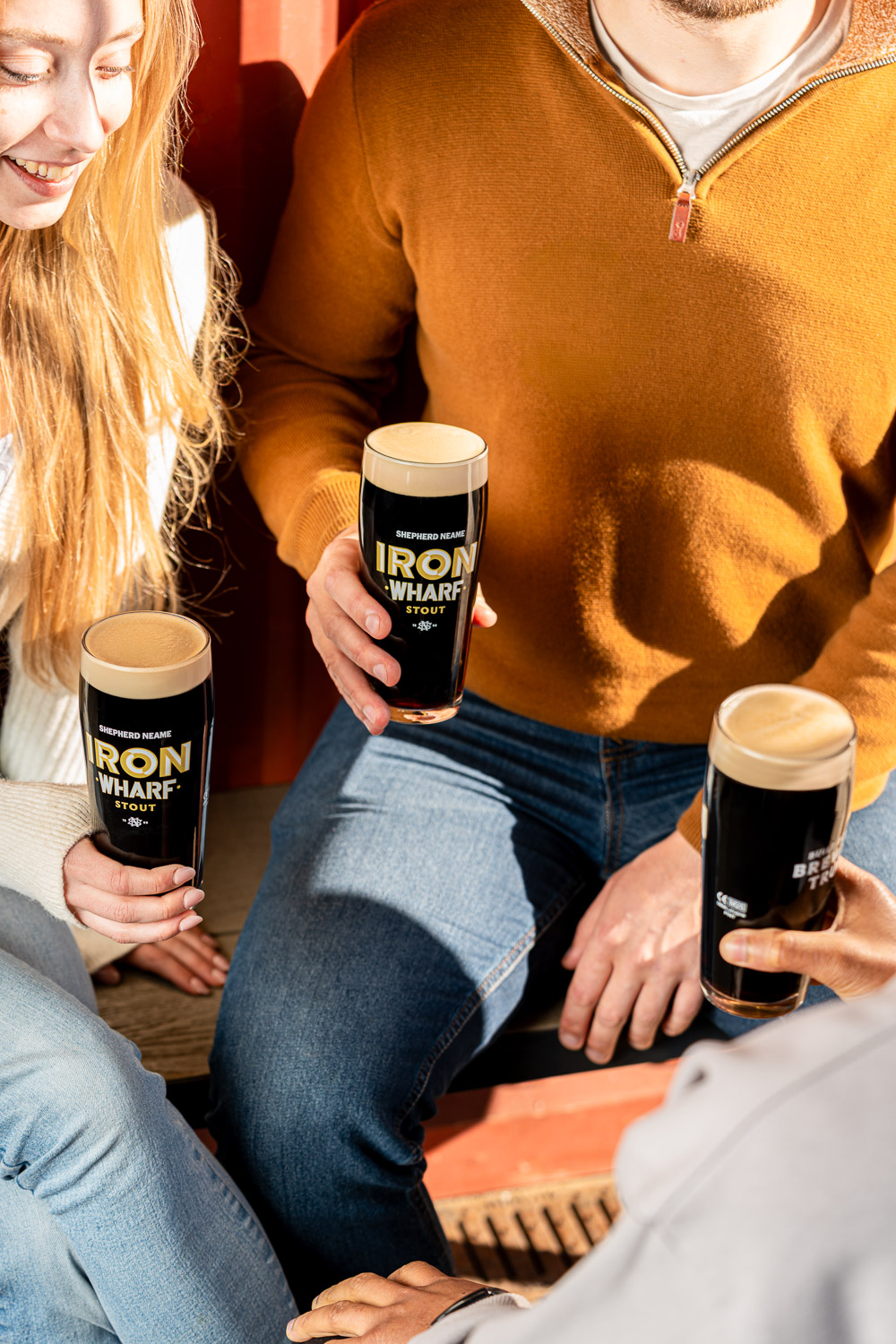 Three glasses of Shepherd Neame's Iron Wharf Stout held by individuals. The dark beer has a thick, light head, and the black glasses feature the white 'Iron Wharf Stout' logo.