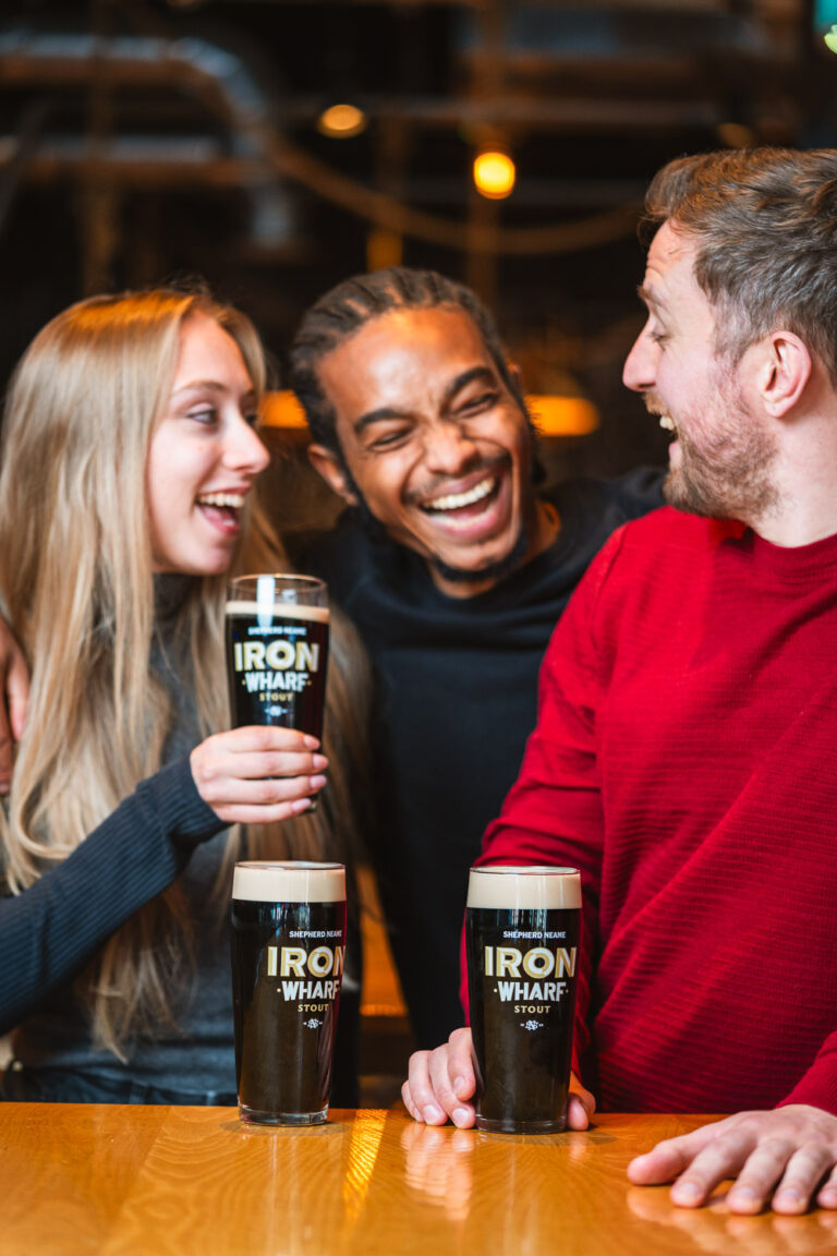 Three friends laughing together while holding pints of Iron Wharf stout at a pub