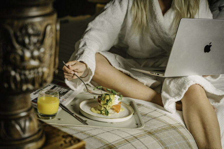 Woman in a robe eating breakfast in bed with orange juice and laptop on her lap