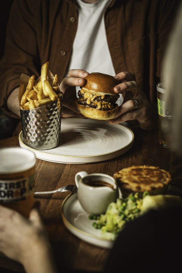 Cheeseburger in brioche bun with fries, pint of beer and side dishes on wooden table