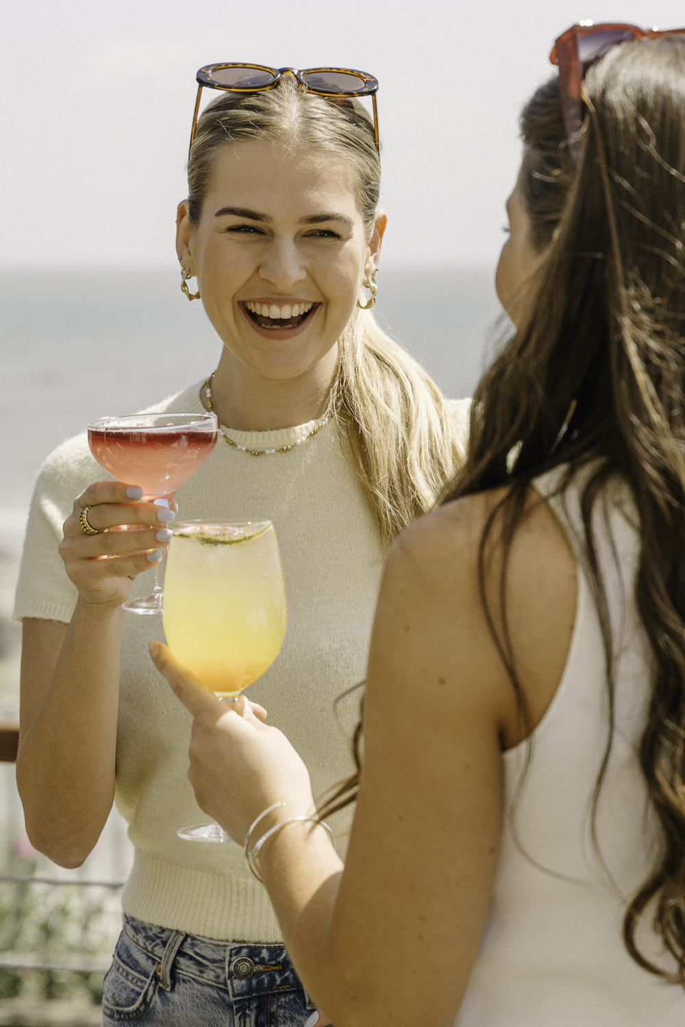 A smiling woman holding a pink cocktail laughs with a friend on a sunny day by the coast. Both women wear sunglasses and light summer outfits, enjoying their colourful drinks outdoors with the sea in the background.
