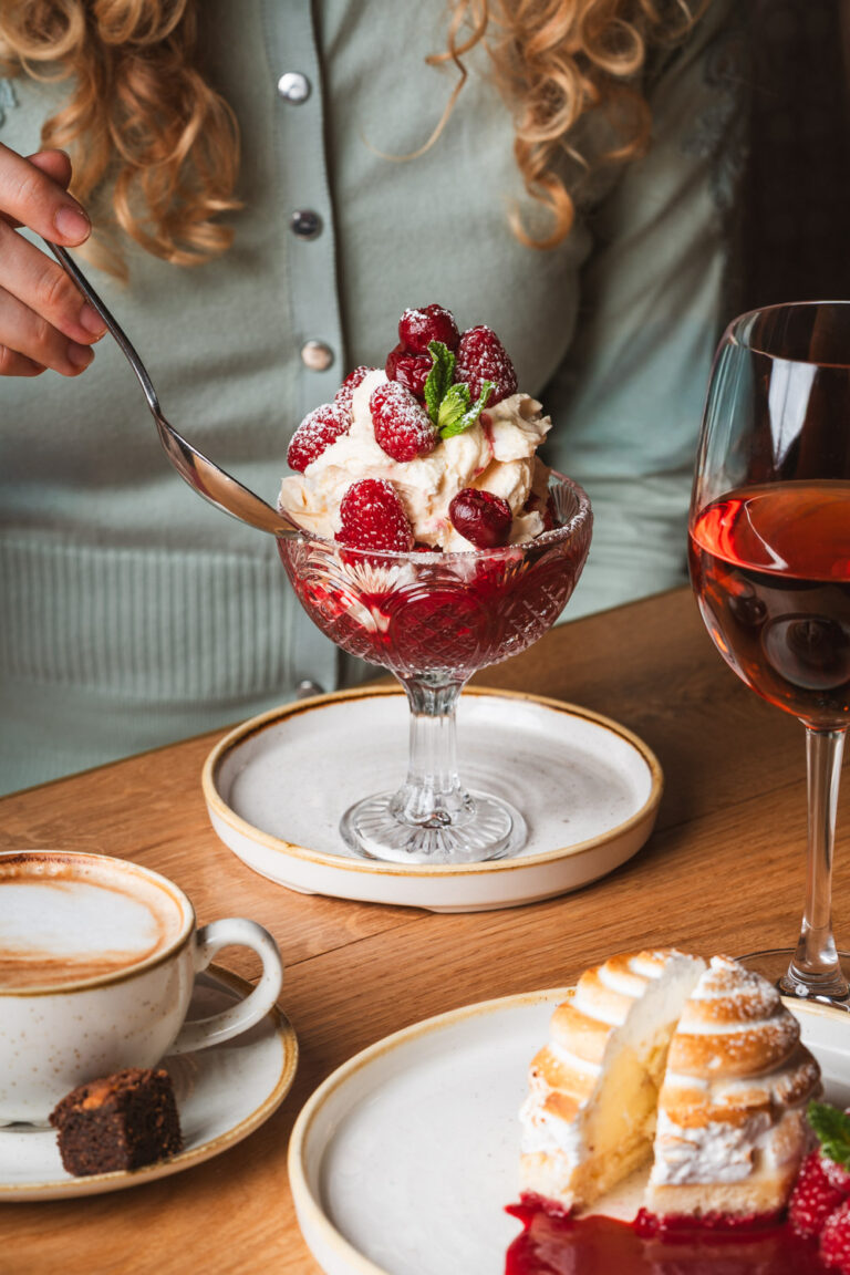 A person holding a spoon about to enjoy a glass dessert bowl filled with whipped cream, raspberries, red currants, and a mint garnish, served on a white plate. On the wooden table are a cappuccino with a brownie bite, a meringue-topped pastry with raspberry sauce, and a glass of rosé wine.