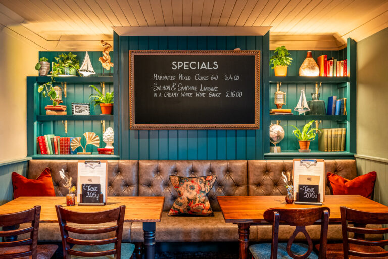 Cozy restaurant interior with a teal-paneled wall, tufted leather bench seating, wooden tables and chairs, and a chalkboard displaying daily specials. The wall features built-in shelves decorated with books, plants, nautical ornaments, and soft ambient lighting.