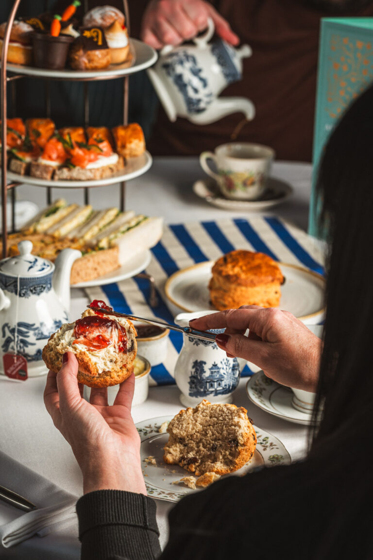Close-up of a traditional afternoon tea setting with a person spreading clotted cream and strawberry jam on a scone. The table features a tiered stand with assorted finger sandwiches, pastries, and cakes, alongside vintage teacups, a blue and white teapot, and a striped tablecloth.