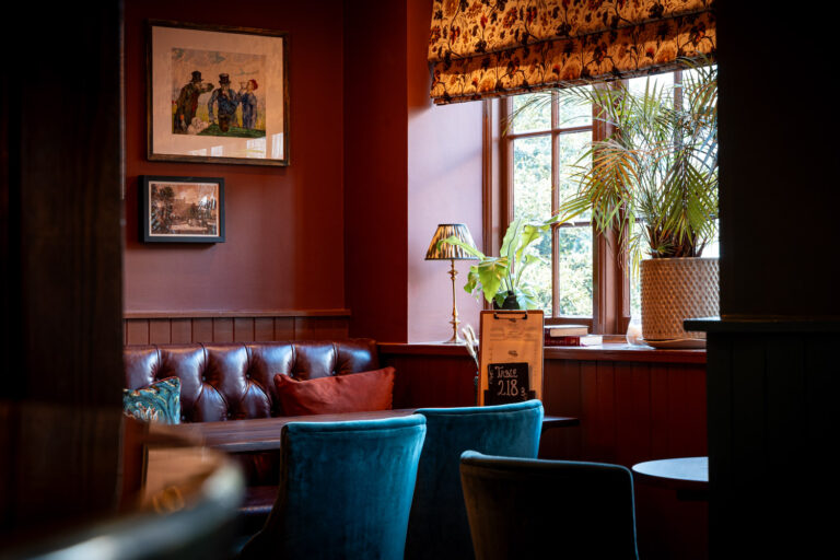 Elegant restaurant seating area with deep red walls, leather bench, blue velvet chairs, and large window with potted plants and floral roman blinds.