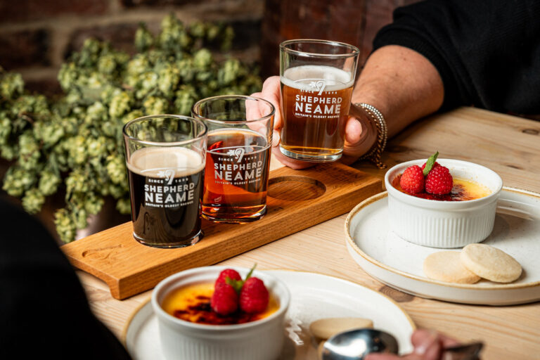 Beer tasting flight with three Shepherd Neame samples on a wooden paddle, a hand holding a fourth glass, next to crème brûlée topped with raspberries and shortbread on a wooden table.