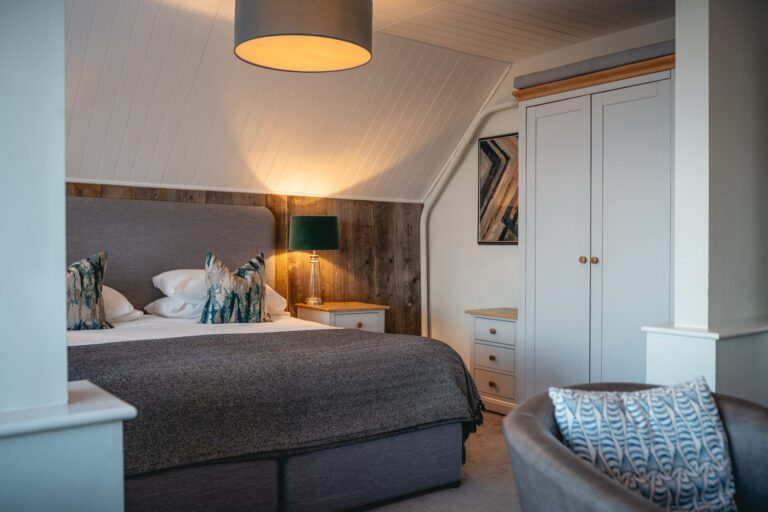 Cozy, modern bedroom with a grey upholstered bed, patterned pillows, and a dark throw blanket, set against a wood-paneled accent wall with a lit table lamp. The room features a white wardrobe, matching bedside tables, and a sloped ceiling with white paneling.