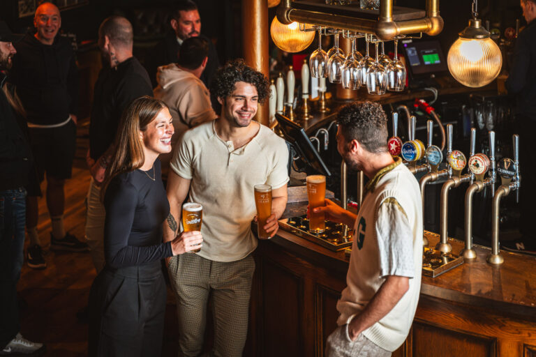 A lively bar scene with three friends smiling and chatting while holding pints of beer at the counter. Behind them, several patrons and bartenders are engaged in conversation. The bar features a row of beer taps and hanging glassware under warm lighting.