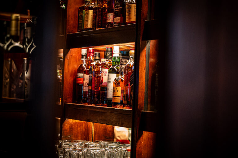 A warmly lit wooden bar shelf stocked with a variety of whiskey and liquor bottles, including brands like Laphroaig and Monkey Shoulder, with glassware neatly arranged on the lower shelf.