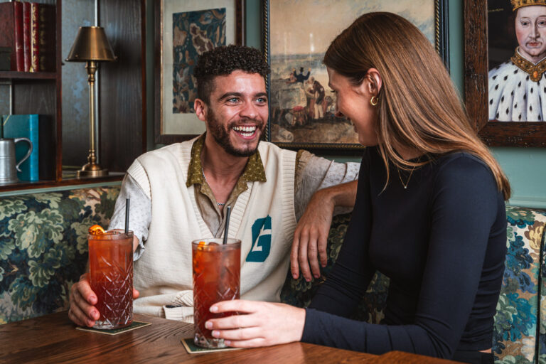 A smiling couple enjoying cocktails in a cozy, vintage-style pub setting. They are seated at a wooden table with two tall iced drinks, surrounded by floral upholstery and framed artwork on the walls.