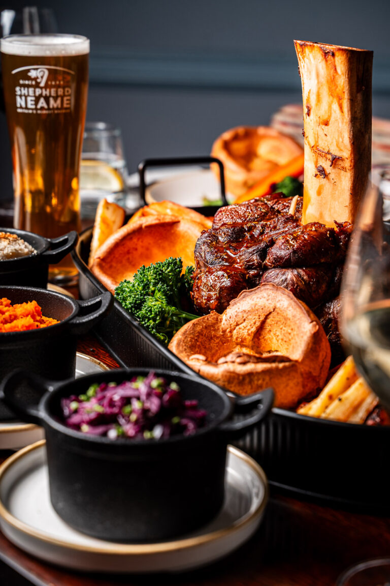A close-up of a traditional Sunday roast platter featuring a large bone-in joint of slow-cooked meat, golden Yorkshire puddings, roasted parsnips, tenderstem broccoli, and vibrant side dishes including braised red cabbage, mashed carrots, and cauliflower cheese in black serving pots. A pint of Shepherd Neame beer and a glass of white wine are visible in the background, enhancing the warm and hearty dining setting.