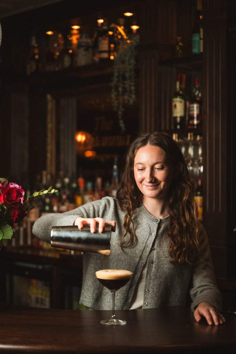Smiling bartender with long wavy hair pours a cocktail from a shaker into a coupe glass at a dimly lit bar, with shelves of liquor bottles and a bouquet of roses in the background.