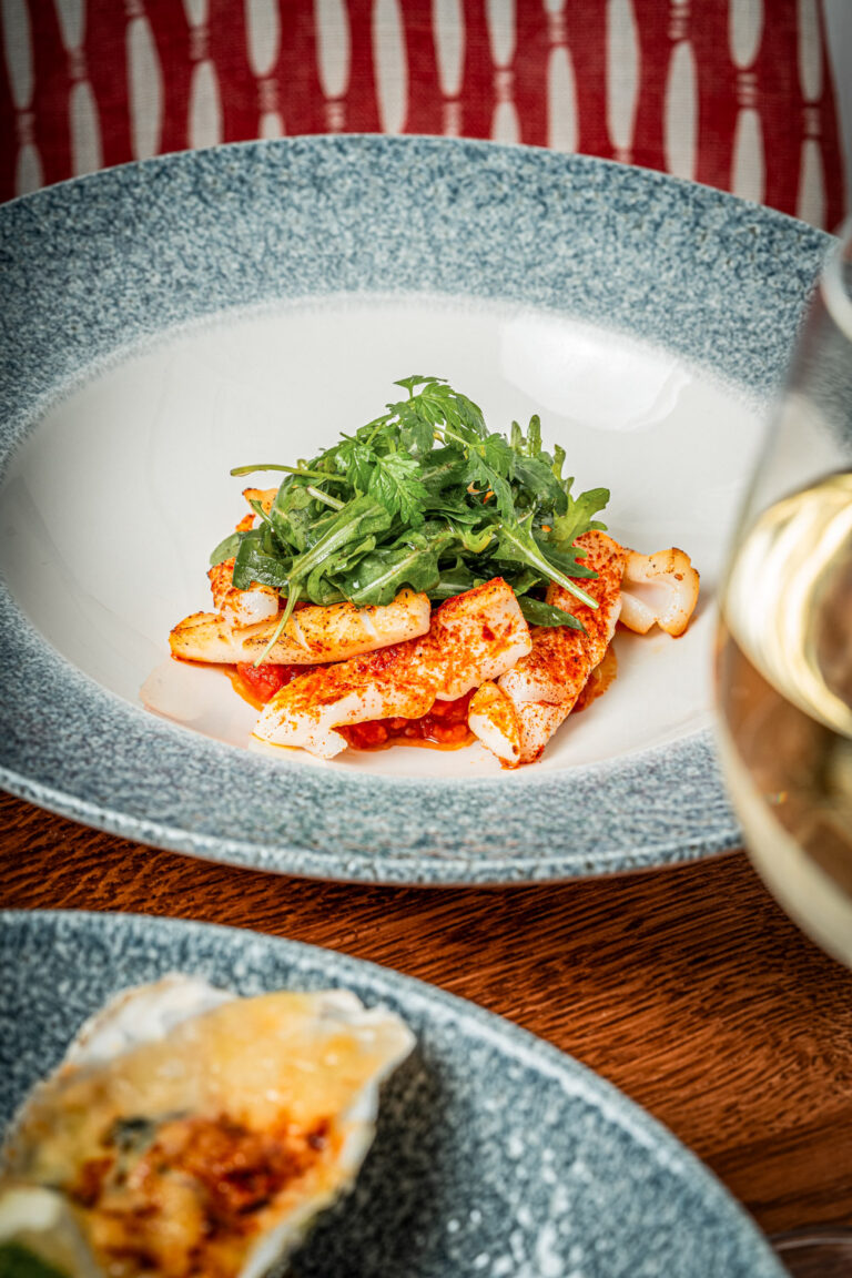 A gourmet dish featuring grilled squid seasoned with spices, served on a bed of tomato sauce and topped with fresh arugula and herbs, presented on a blue-rimmed plate. A blurred glass of white wine and another plated dish appear in the foreground.