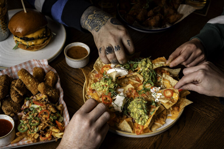 Close-up of friends sharing nachos topped with guacamole, sour cream, cheese, and chillies at The Crown pub in Blackheath, with burgers and sharing plates on the table.