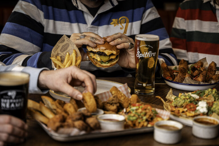 Friends sharing a table of burgers, fries, chicken wings, nachos, and pints of Spitfire beer at The Crown pub in Blackheath.
