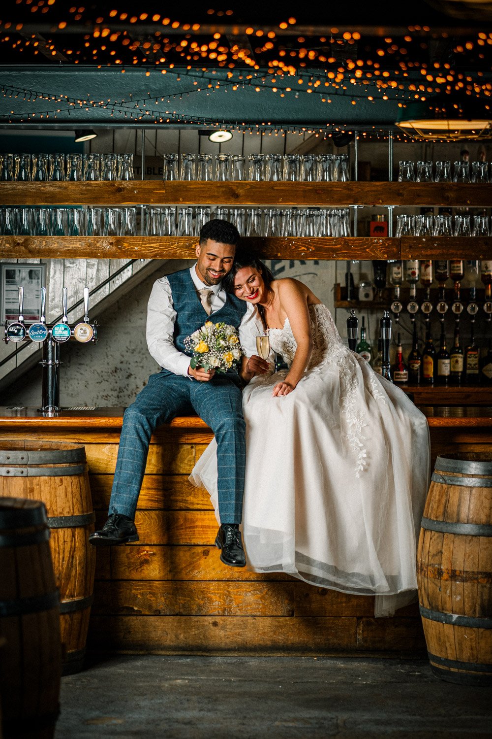 Bride and groom sitting on a rustic wooden bar counter, smiling and holding a bouquet and champagne glass, with fairy lights above and shelves of glassware behind them in a cozy, warmly lit bar setting.