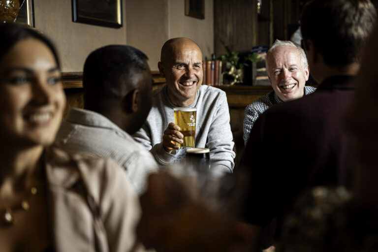 Group of friends enjoying drinks and conversation inside The White Horse & Bower pub, with a smiling man holding a pint of beer.