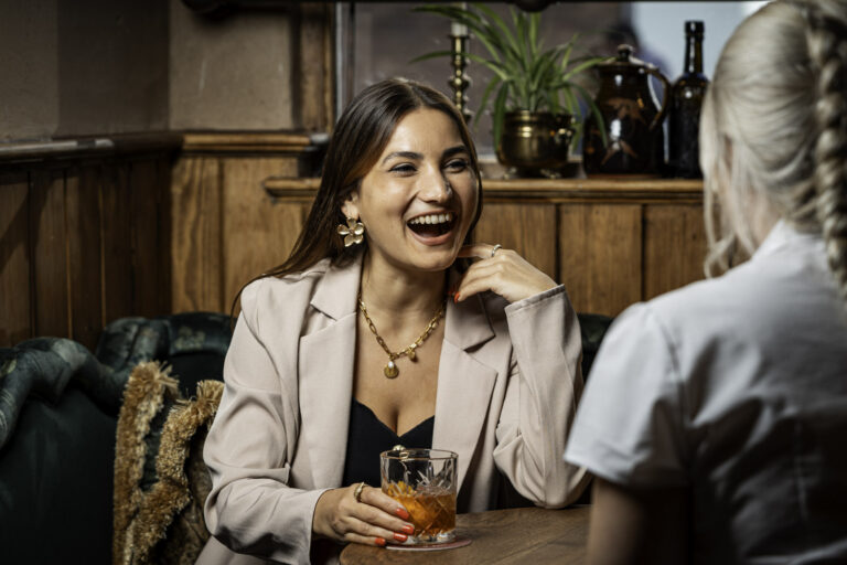 Woman smiling and holding a cocktail while chatting with a friend inside The White Horse & Bower pub in London.