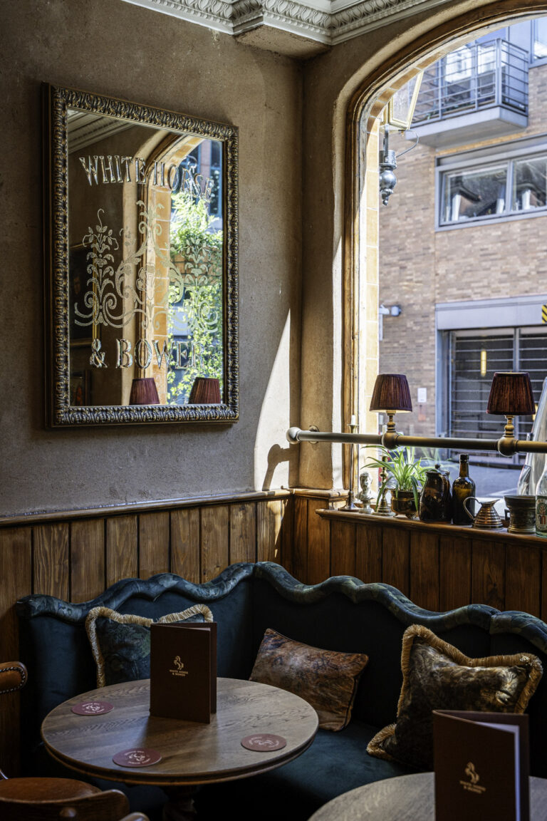 Cozy pub corner at The White Horse & Bower with a vintage mirror, green velvet seating, wooden tables, and soft natural light.