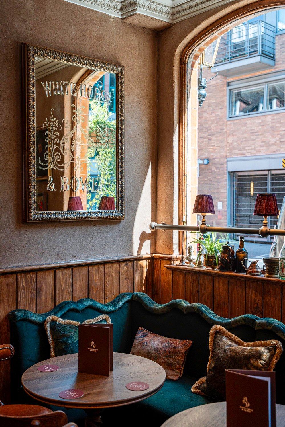 A welcoming spot inside The White Horse & Bower. Cozy pub corner at The White Horse & Bower with a vintage mirror, green velvet seating, wooden tables, and soft natural light.