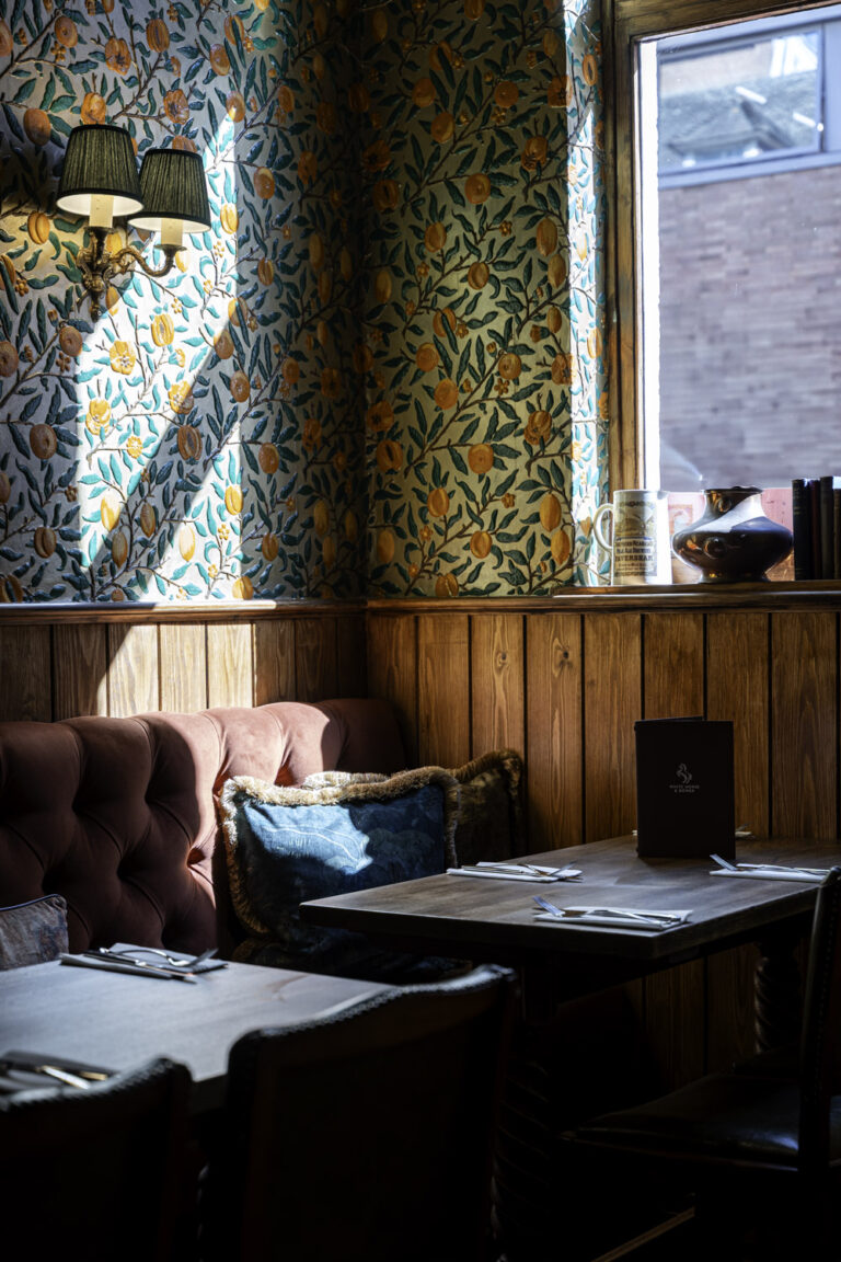 Intimate pub seating area with a wooden table, cushioned chairs, floral wallpaper, and natural light streaming through the window.