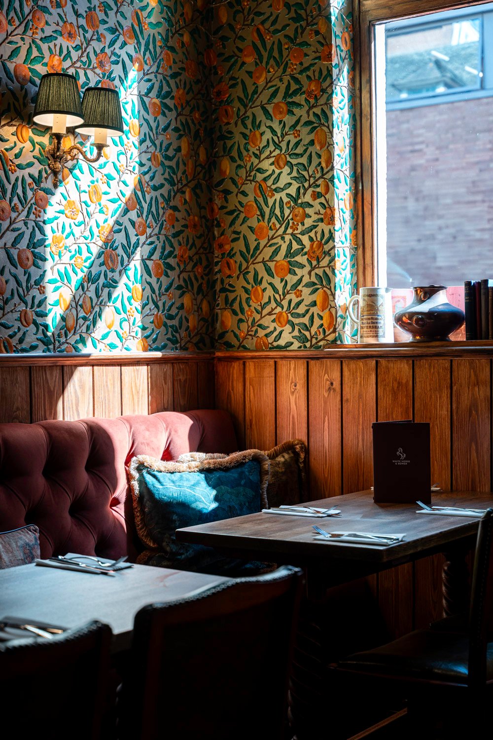 Relax in style at The White Horse & Bower. Intimate pub seating area with a wooden table, cushioned chairs, floral wallpaper, and natural light streaming through the window.