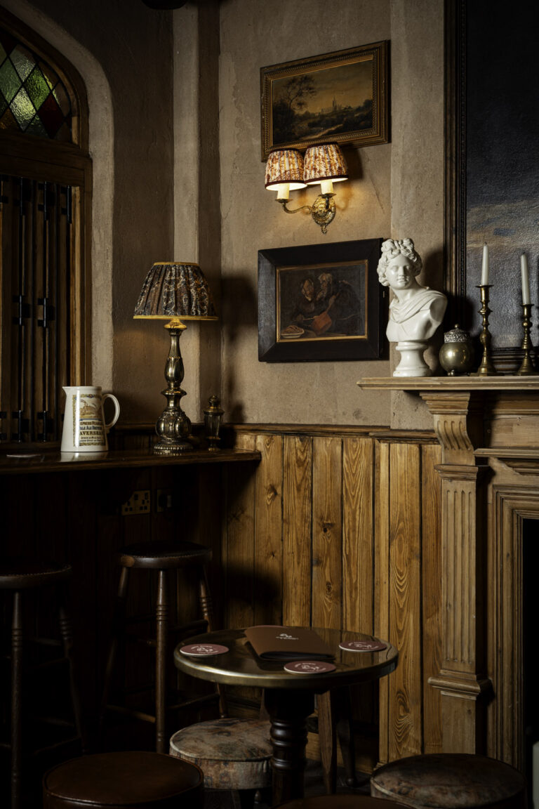 Traditional pub interior with wooden paneling, a round table with coasters, leather stools, vintage lamps, framed artwork, and a white bust on the mantel.