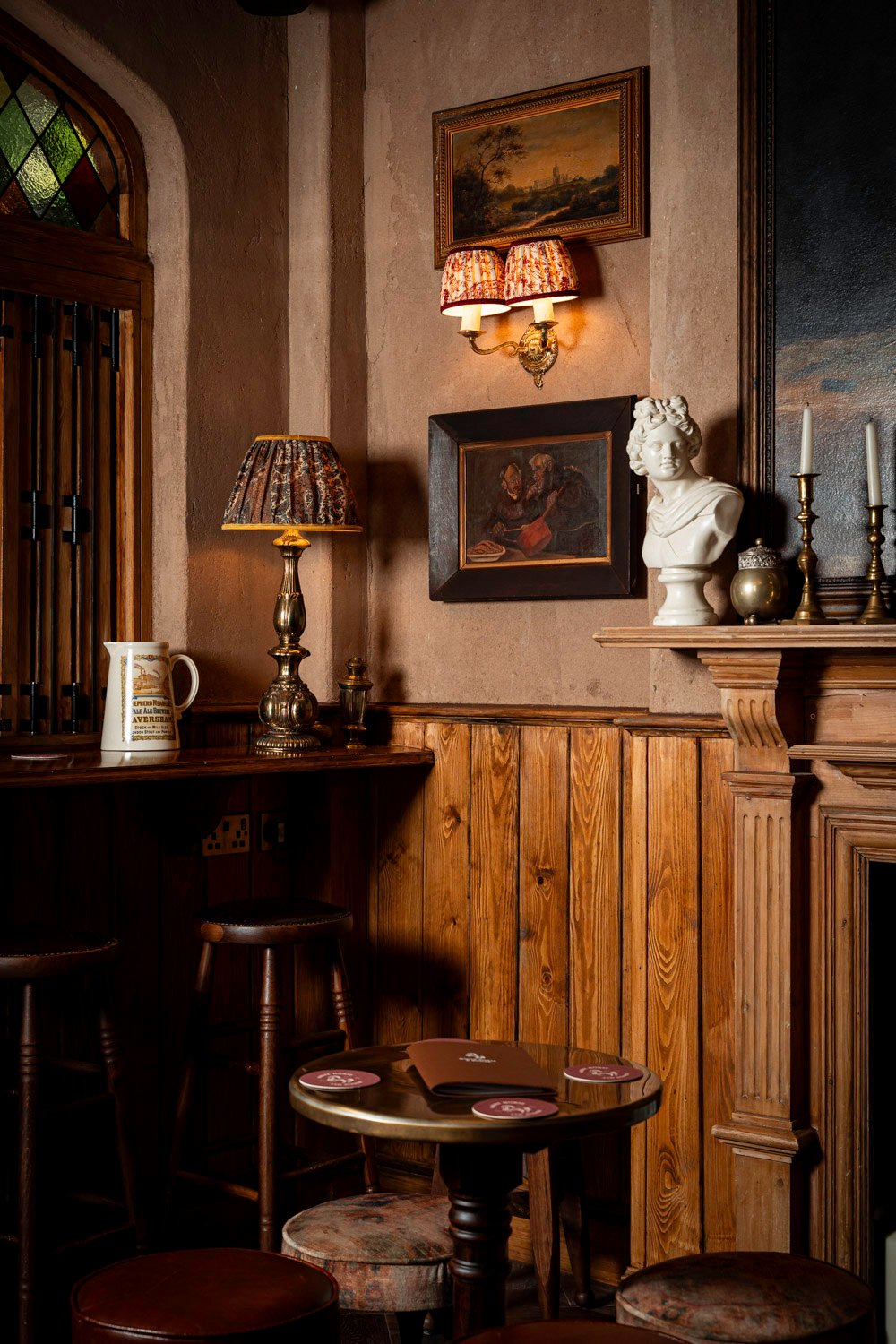 Classic pub charm inside The White Horse & Bower. Traditional pub interior with wooden paneling, a round table with coasters, leather stools, vintage lamps, framed artwork, and a white bust on the mantel.