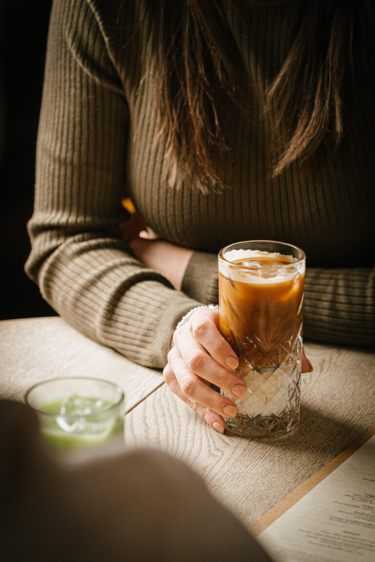A person wearing a ribbed olive green sweater is holding a glass of iced coffee with both hands on a rustic wooden table. Another blurry glass, containing a green beverage, is also on the table.