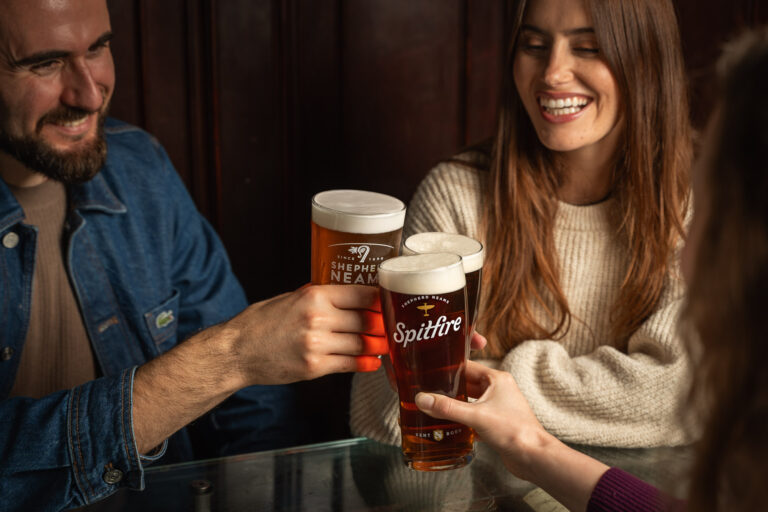 Three friends clinking pints of amber beer labeled “Shepherd Neame” and “Spitfire” while smiling and sitting together at a pub table.