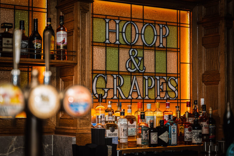 Stained glass Hoop & Grapes sign behind a traditional London pub bar with illuminated spirits display.