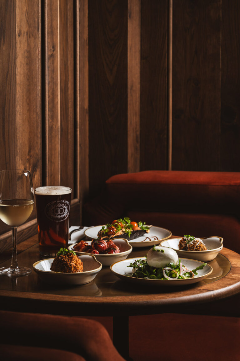 Small plates and tapas dishes with wine and beer on a wooden table in a moody restaurant setting with warm lighting.