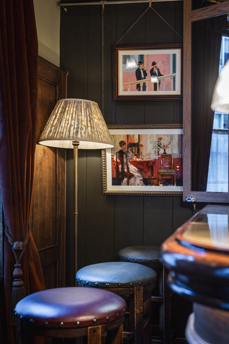 Traditional London pub interior with warm lamp lighting, wooden panelling, framed artwork and bar stools.