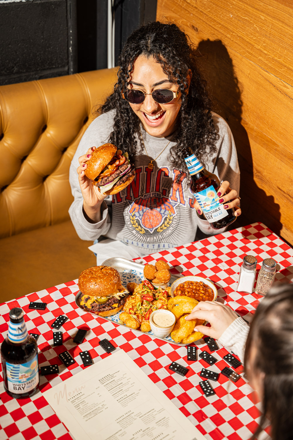 Professional food and lifestyle photography for restaurants, bars, and hospitality brands. We create compelling visual content that drives engagement and showcases your venue's atmosphere and menu. Customer holding burger and beer at casual dining restaurant with retro booth seating and red checkered tablecloth