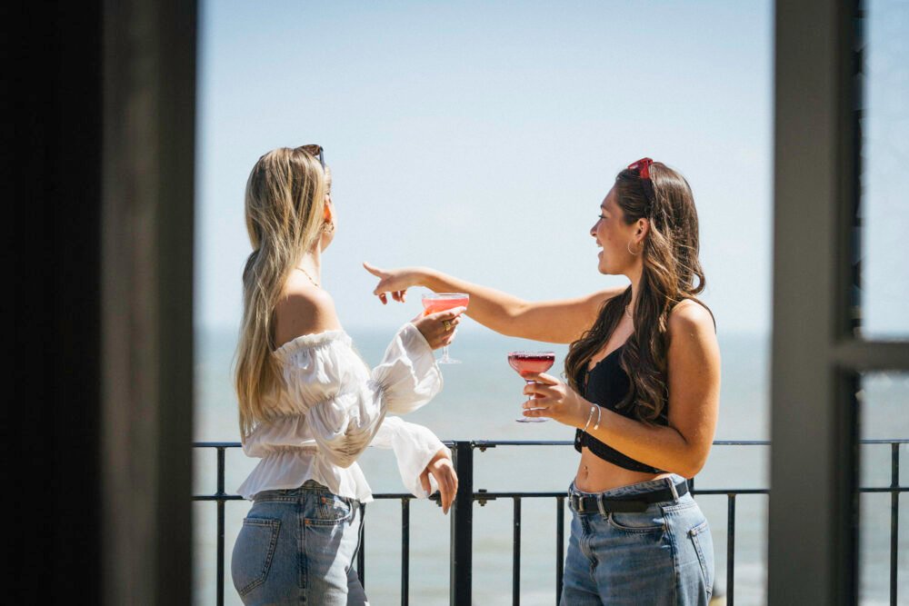 Two women with cocktails pointing out to sea on a sunny coastal balcony