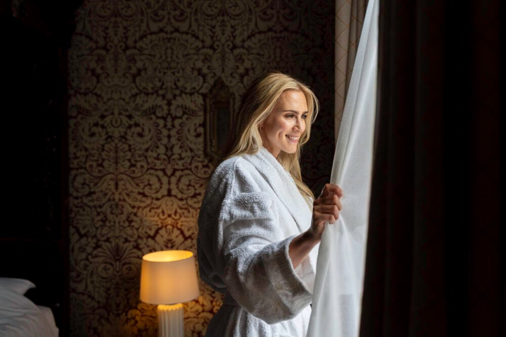 A smiling woman in a white bathrobe stands by a window, gently pulling back a sheer curtain as soft daylight illuminates her face. The room features ornate, vintage wallpaper and a glowing bedside lamp, creating a warm and elegant atmosphere.