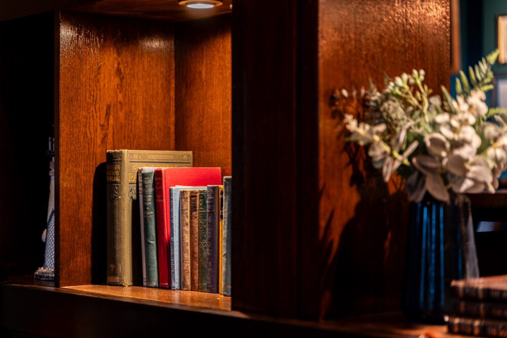 A cozy corner with antique books and floral decor. Close-up of a wooden bookshelf with vintage books under warm spotlight, next to a vase of white flowers.