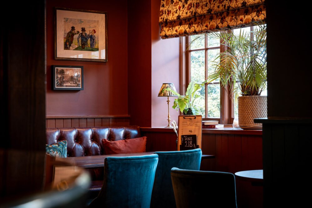 Stylish and intimate dining nook with natural light and classic decor. Elegant restaurant seating area with deep red walls, leather bench, blue velvet chairs, and large window with potted plants and floral roman blinds.