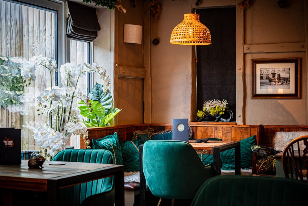 Cozy café interior with teal velvet chairs, wooden tables, rattan pendant light, white orchids by a window, and a framed vintage photo above cushioned seating.