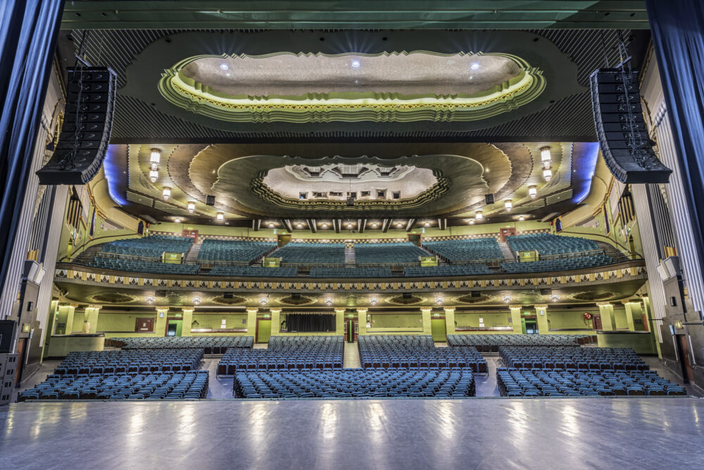 Inside the Eventim Apollo (formerly the Hammersmith Odeon) in Hammersmith, London. View from the stage inside the Eventim Apollo in Hammersmith, London — showing tiered turquoise seating and an ornate Art Deco ceiling.