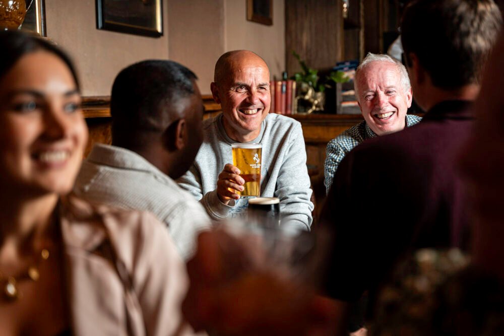 Enjoy the friendly atmosphere of The White Horse & Bower, a Shepherd Neame pub in London. Perfect for meeting friends, sharing laughs, and enjoying a pint in a classic British pub setting. Group of friends enjoying drinks and conversation inside The White Horse & Bower pub, with a smiling man holding a pint of beer.