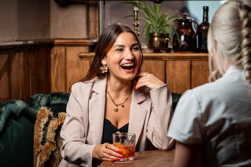 Cocktails, laughter, and good company at The White Horse & Bower. Woman smiling and holding a cocktail while chatting with a friend inside The White Horse & Bower pub in London.