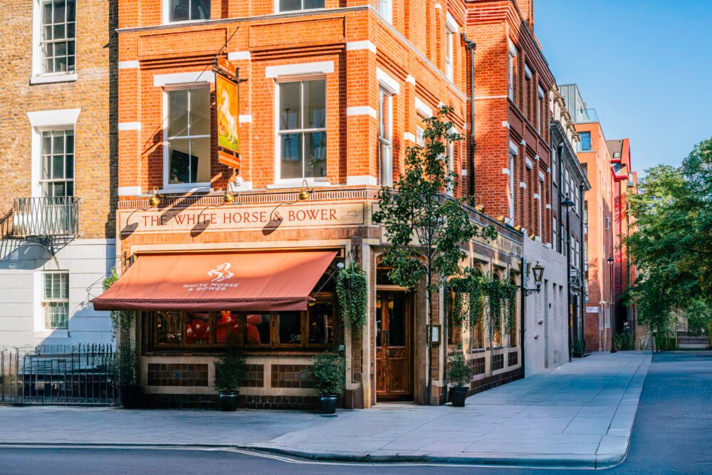The White Horse & Bower – a traditional Shepherd Neame pub on a quiet London street Street view of The White Horse & Bower pub in London, a red-brick building with large windows, hanging greenery, and a red canopy sign.