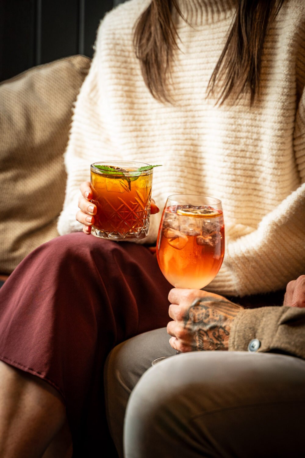 Two people holding iced cocktails in tumbler glasses, warm cosy setting