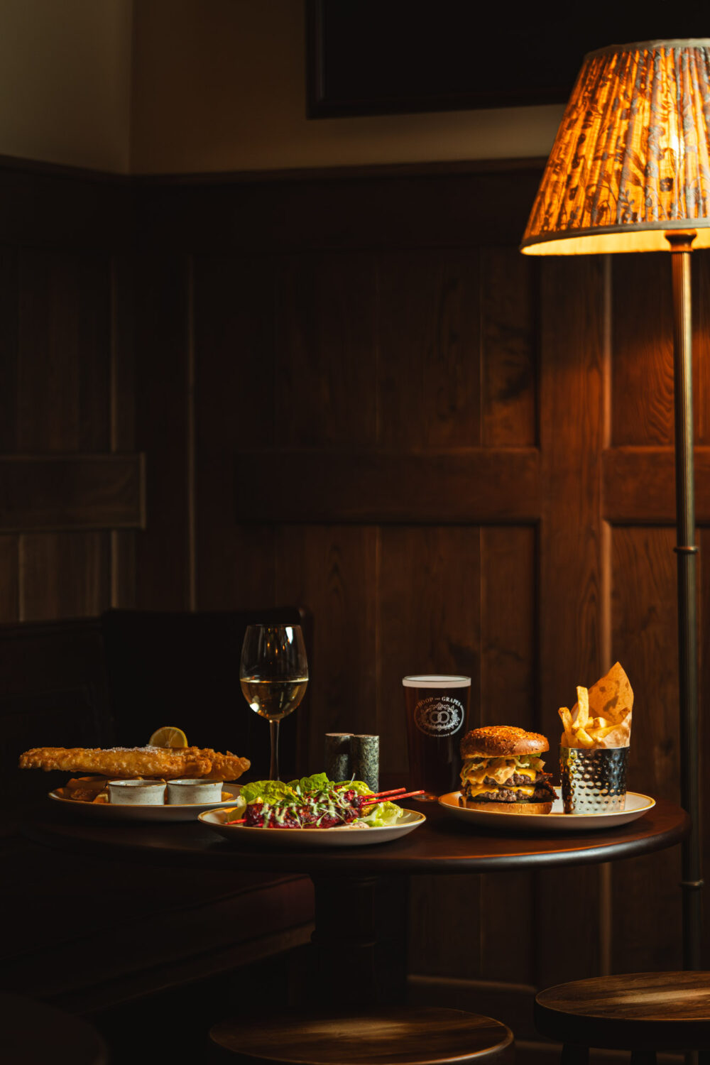 A cosy gastropub scene with fish and chips, stacked burger and fries, fresh salad and wine under warm ambient lighting. Fish and chips, gourmet burger with fries, salad and wine on a table in a cosy gastropub with warm ambient lighting.