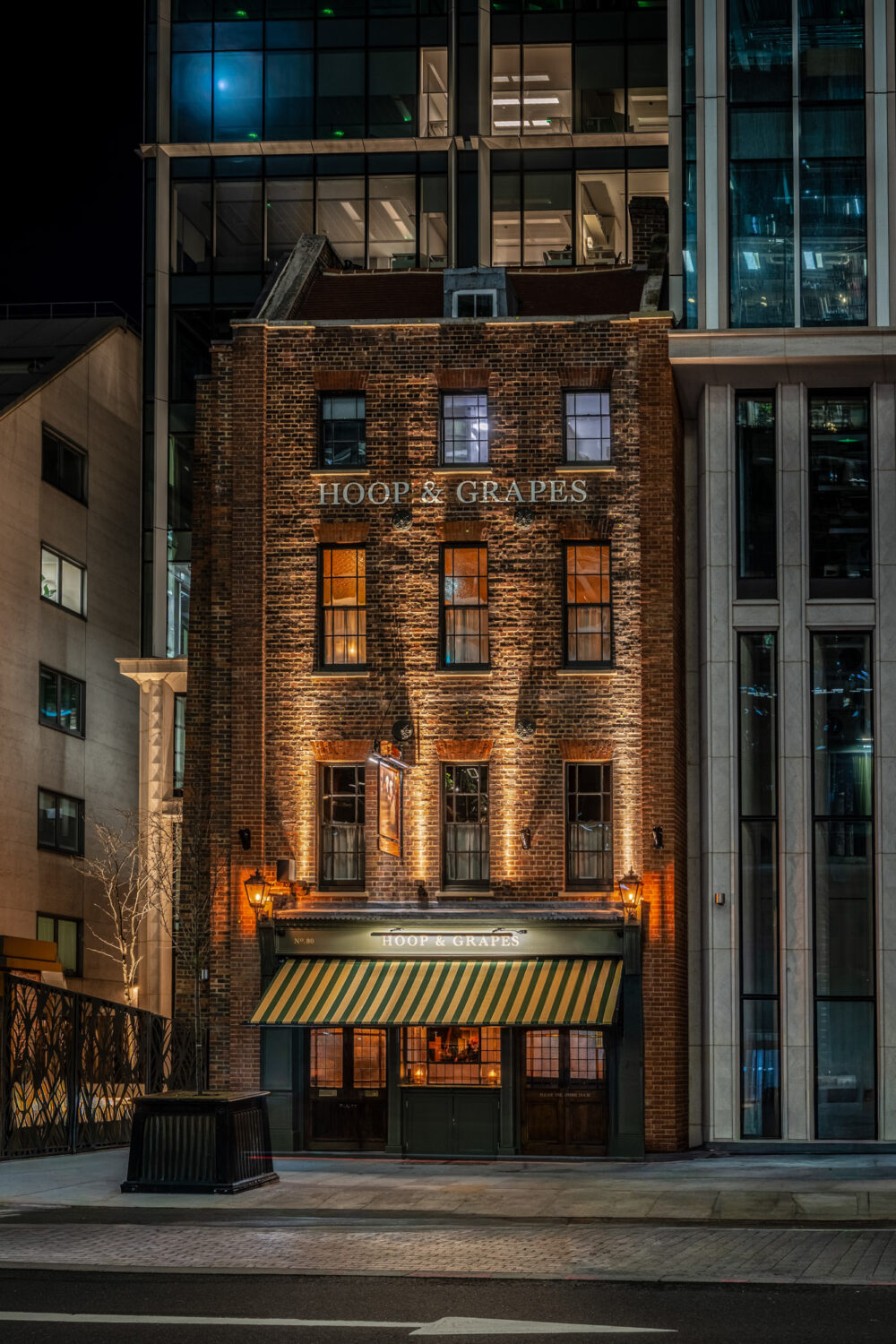 The historic Hoop & Grapes pub in London captured at night, glowing warmly against the surrounding modern architecture. Hoop & Grapes historic London pub exterior at night, warmly lit brick building surrounded by modern city architecture.