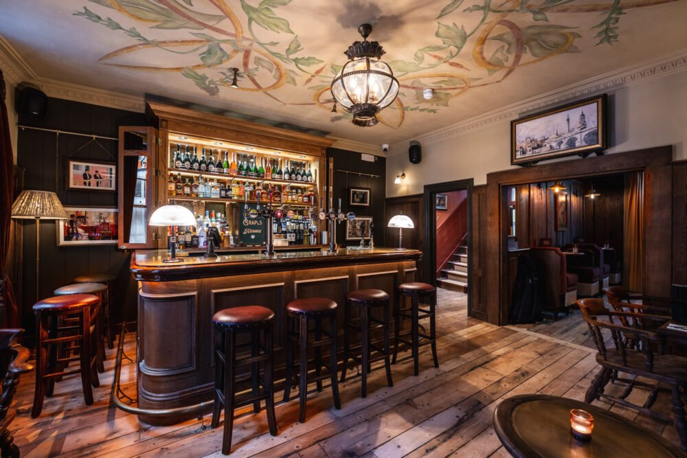 The characterful bar at a traditional London pub, captured with warm lighting and rich wood textures. Traditional London pub bar interior with wooden panelling, bar stools, spirits display and warm ambient lighting.